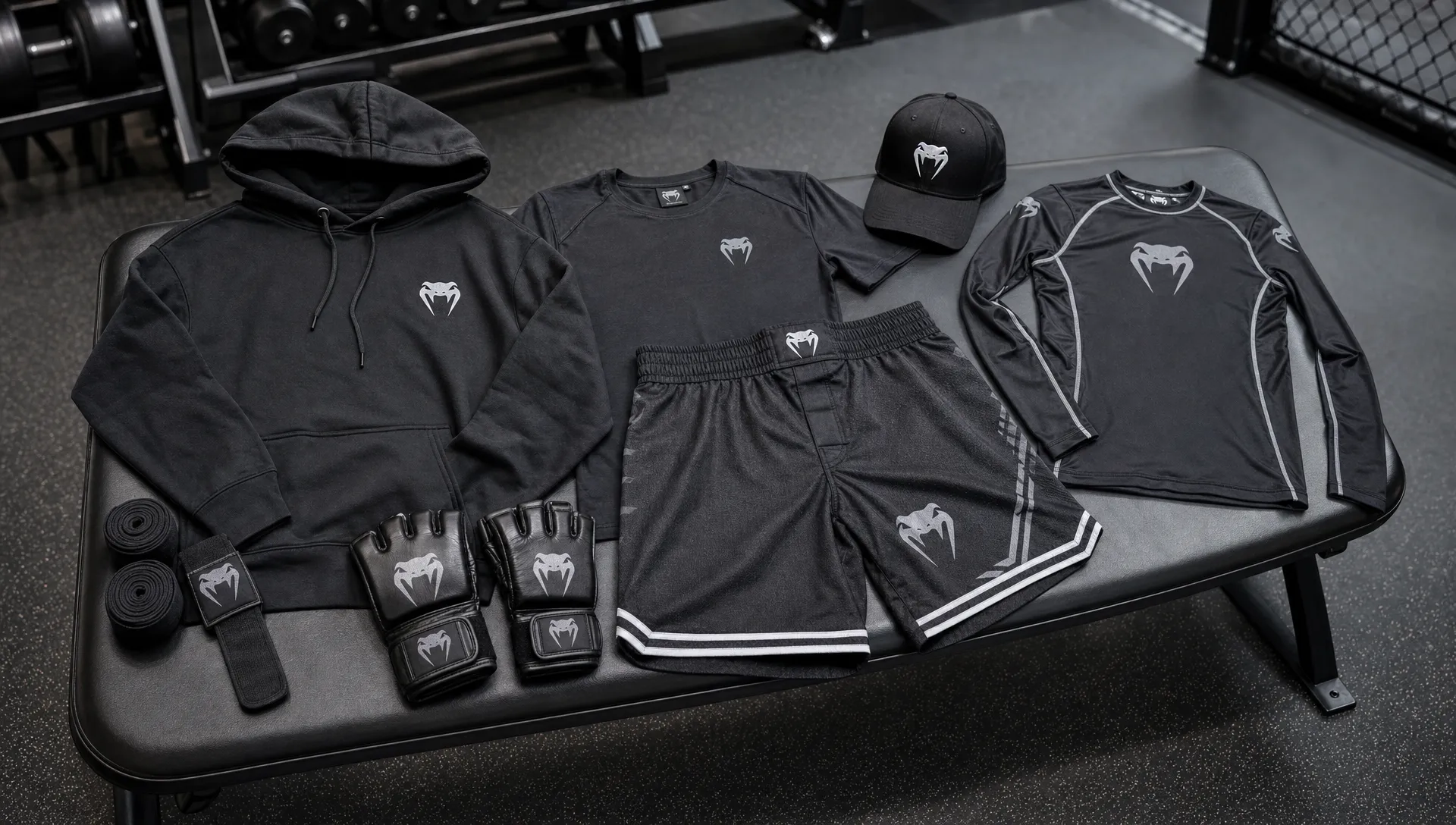 A gym bench holds a premium hoodie, performance tee, fight shorts, cap, rash guard, hand wraps, and MMA gloves arranged as lifestyle MMA merch.