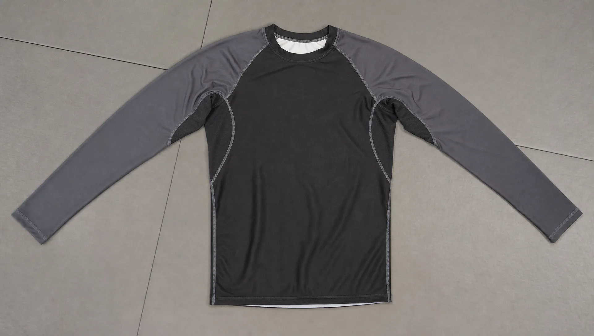 A close view of a martial arts rash guard laid flat on a training mat, with the neckline, raglan shoulder seams, underarm gusset panels, side seams, and hem visible.