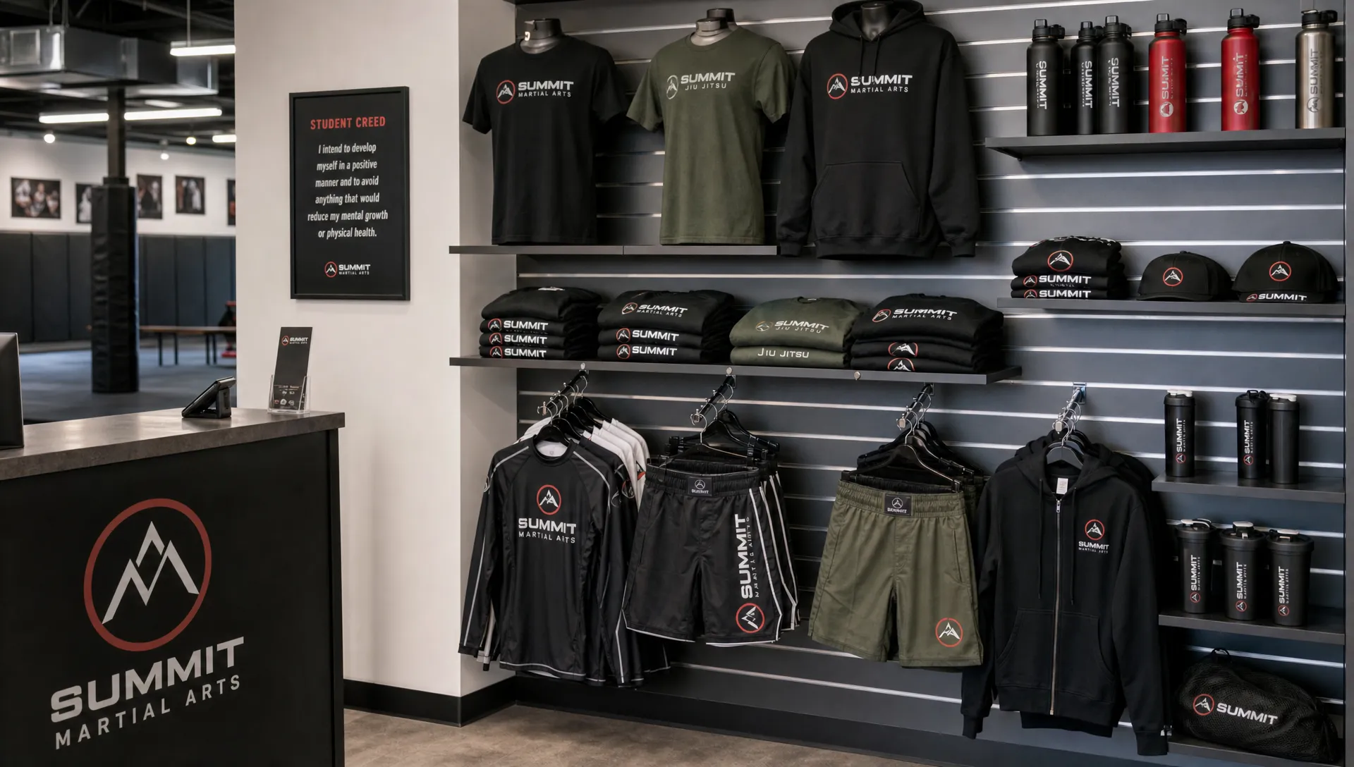 A martial arts gym retail wall shows folded branded shirts, hanging rash guards, training shorts, hoodies, and water bottles near reception.