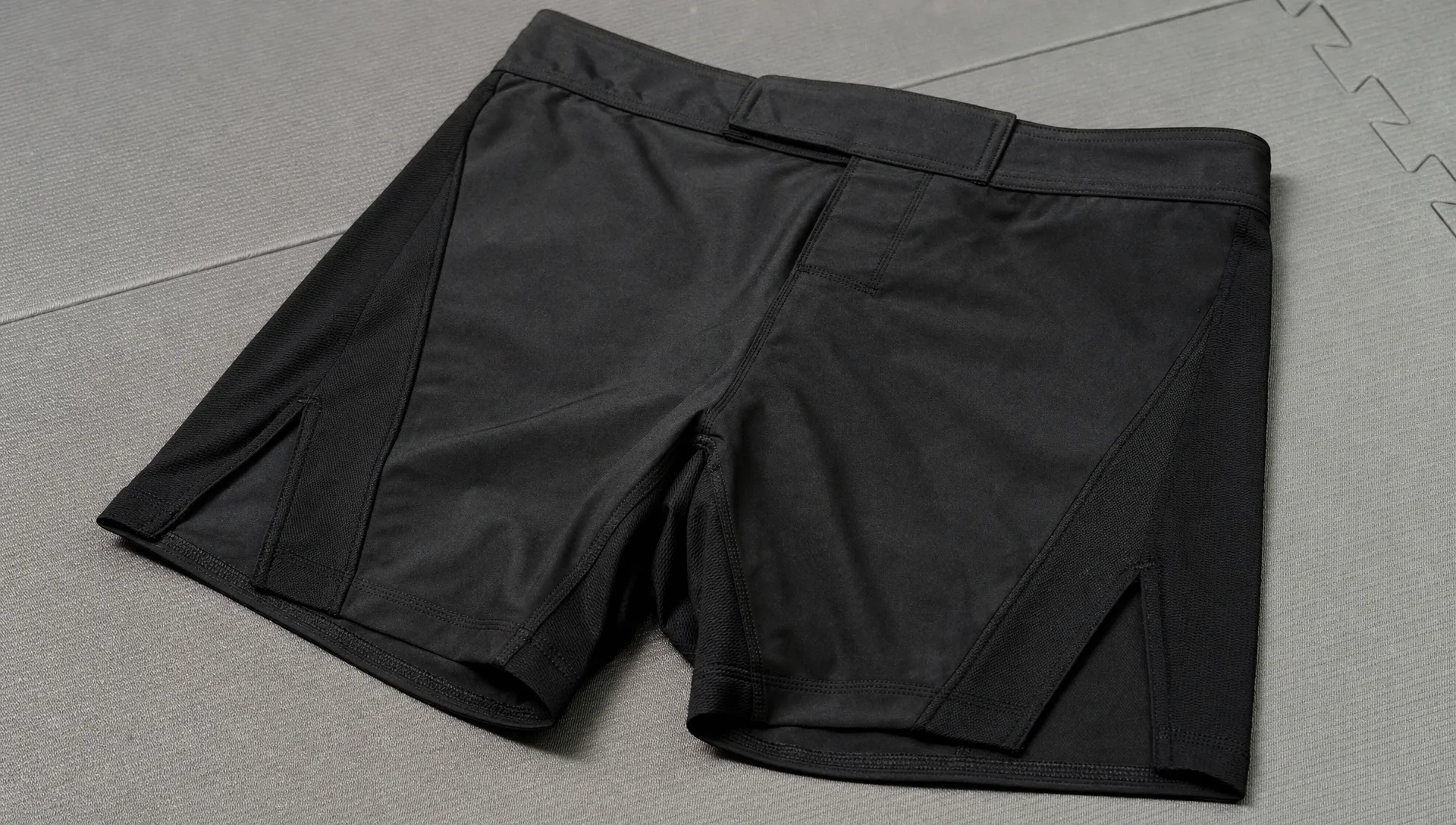 Close-up of black BJJ fight shorts on a grappling mat, showing the flat waistband, stretch panels and reinforced seams used for no-gi training.