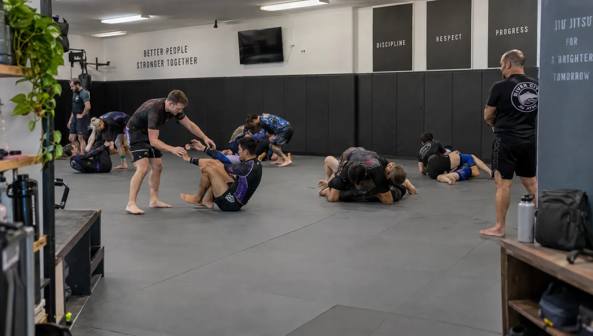 Adult athletes in rash guards drill grappling moves across a busy gym mat as a coach watches nearby.