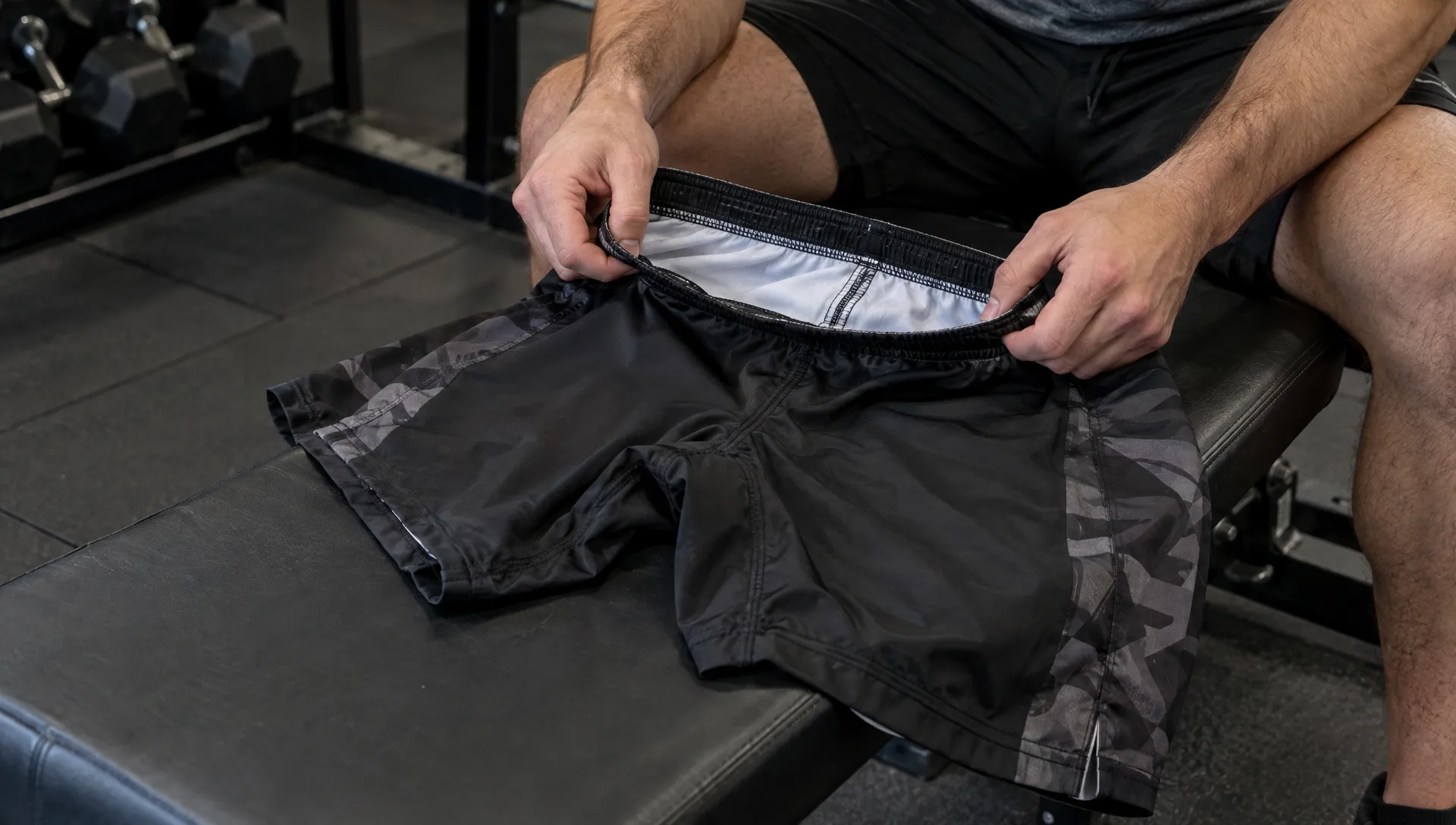 BJJ shorts are stretched across a gym bench as a tester checks the waistband interior and crotch seams after a movement session.