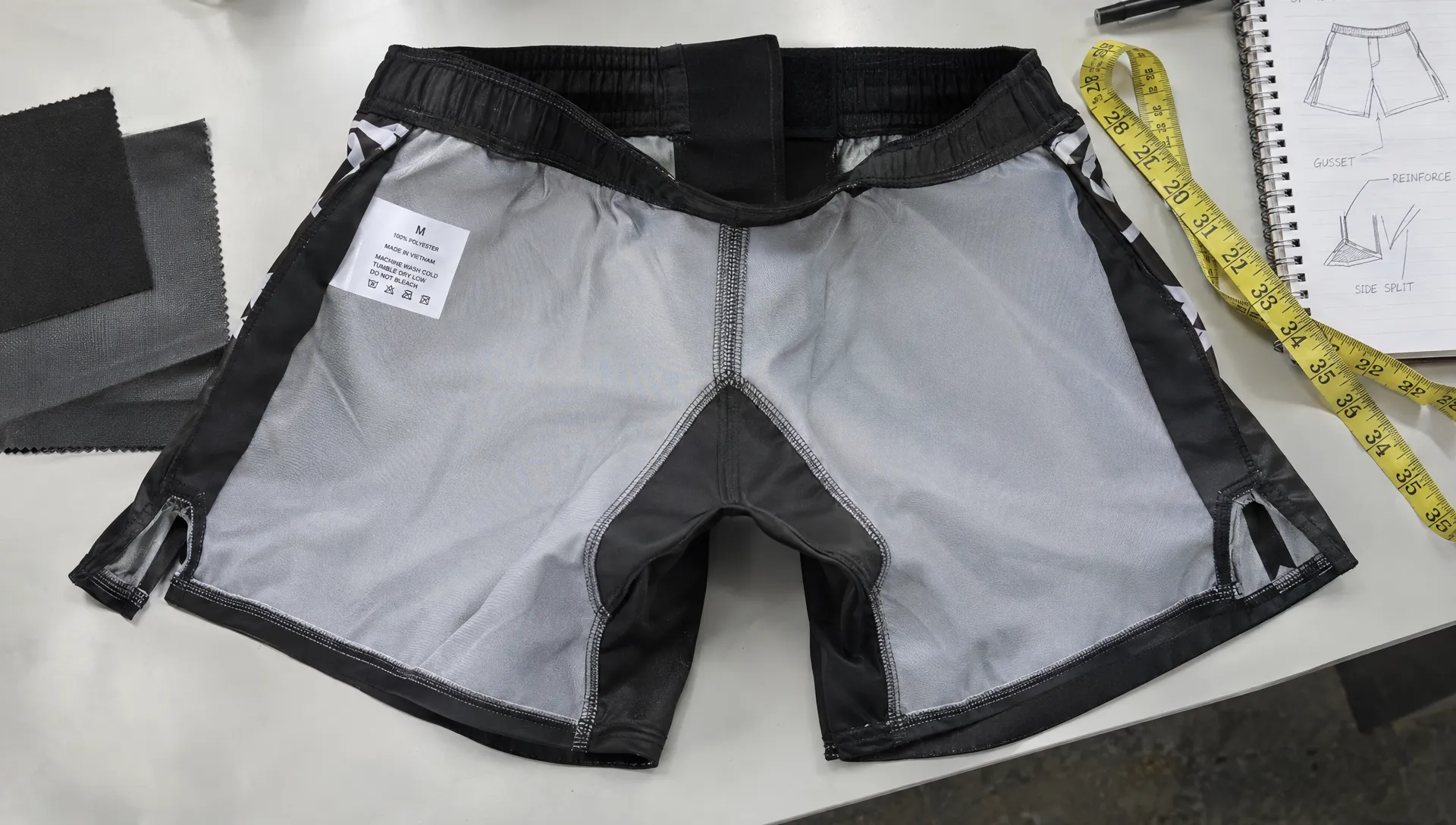Overhead view of fightwear shorts opened inside out on a worktable, with the gusset, covered closure, smooth seams and reinforced side-split ends visible beside a fabric swatch and measuring tape.